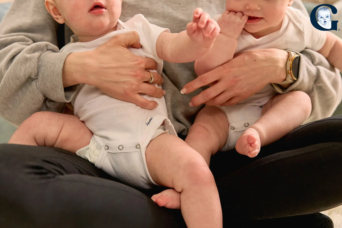 Two babies in white short-sleeve Gerber bodysuits sitting on a parent's lap