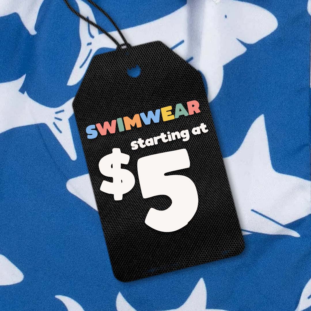 A black fabric price tag hanging from a black string, printed with the text 'SWIMWEAR starting at $5' in multicolor and white lettering, placed on a blue and white shark-patterned garment.
