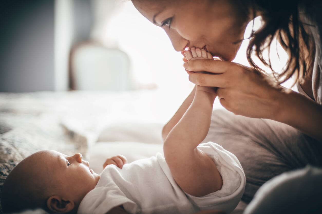 Loving mother kissing newborn baby’s tiny feet while the baby rests in a soft white Gerber Childrenswear Onesies® bodysuit. This intimate moment captures the comfort and softness of Gerber baby clothes, designed for precious newborn moments. Shop Gerber Childrenswear for affordable newborn essentials, baby layette sets, and everyday baby clothing deals.