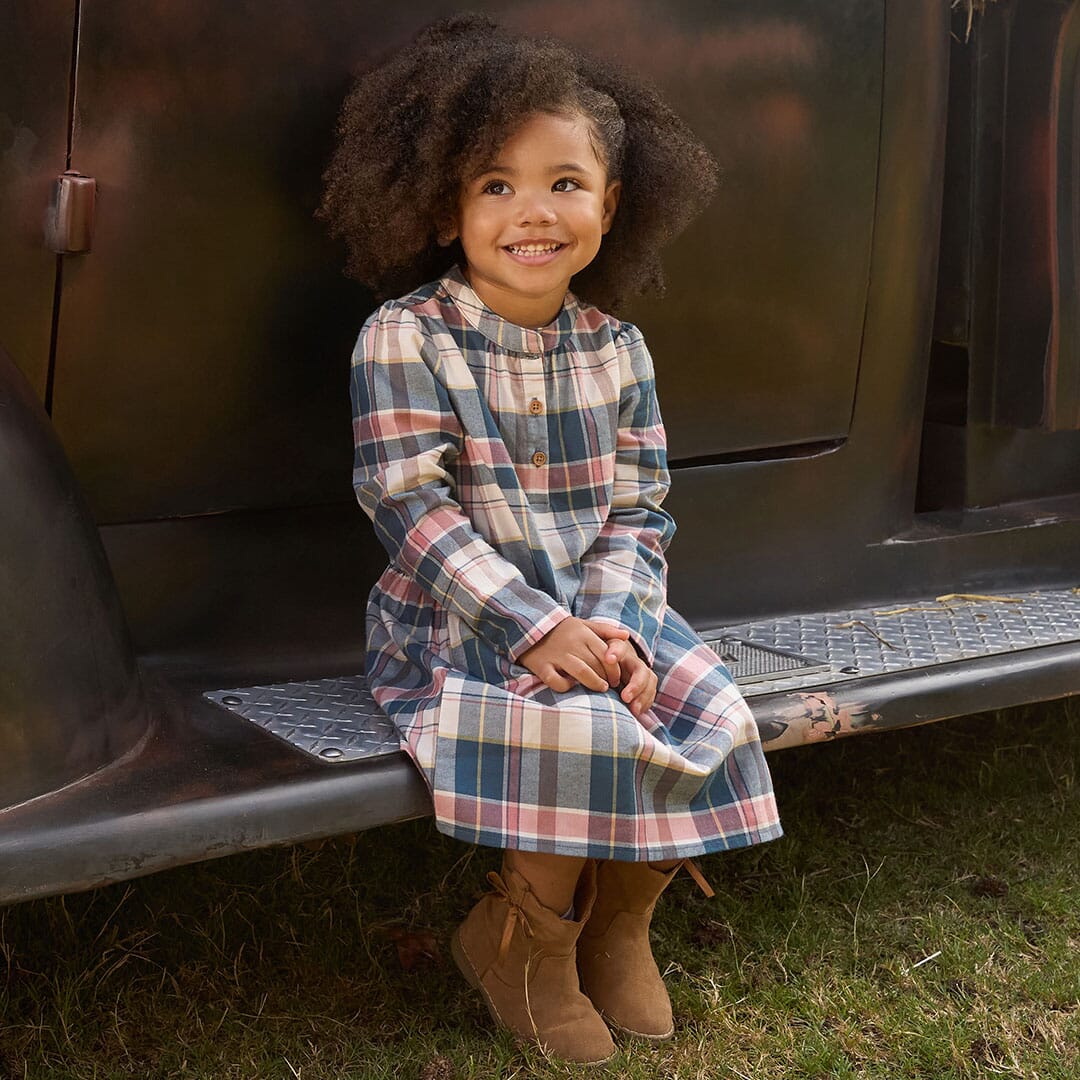 Child wearing a plaid dress sitting on a vehicle bumper outdoors