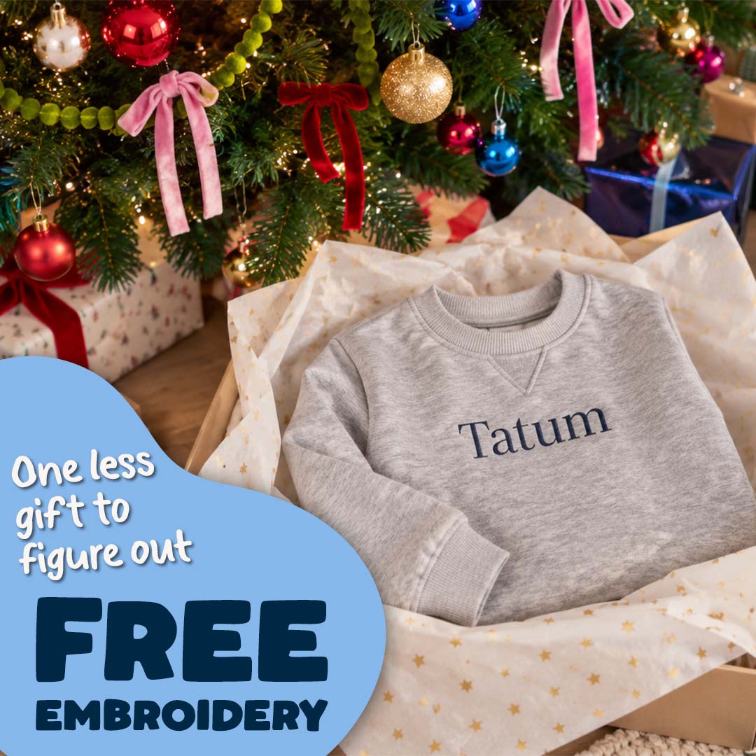 A light heather gray long-sleeve sweatshirt with the name 'Tatum' embroidered in navy on the chest, presented in a gift box lined with tissue paper under a decorated Christmas tree. A blue promotional overlay on the left reads 'One less gift to figure out FREE EMBROIDERY'.