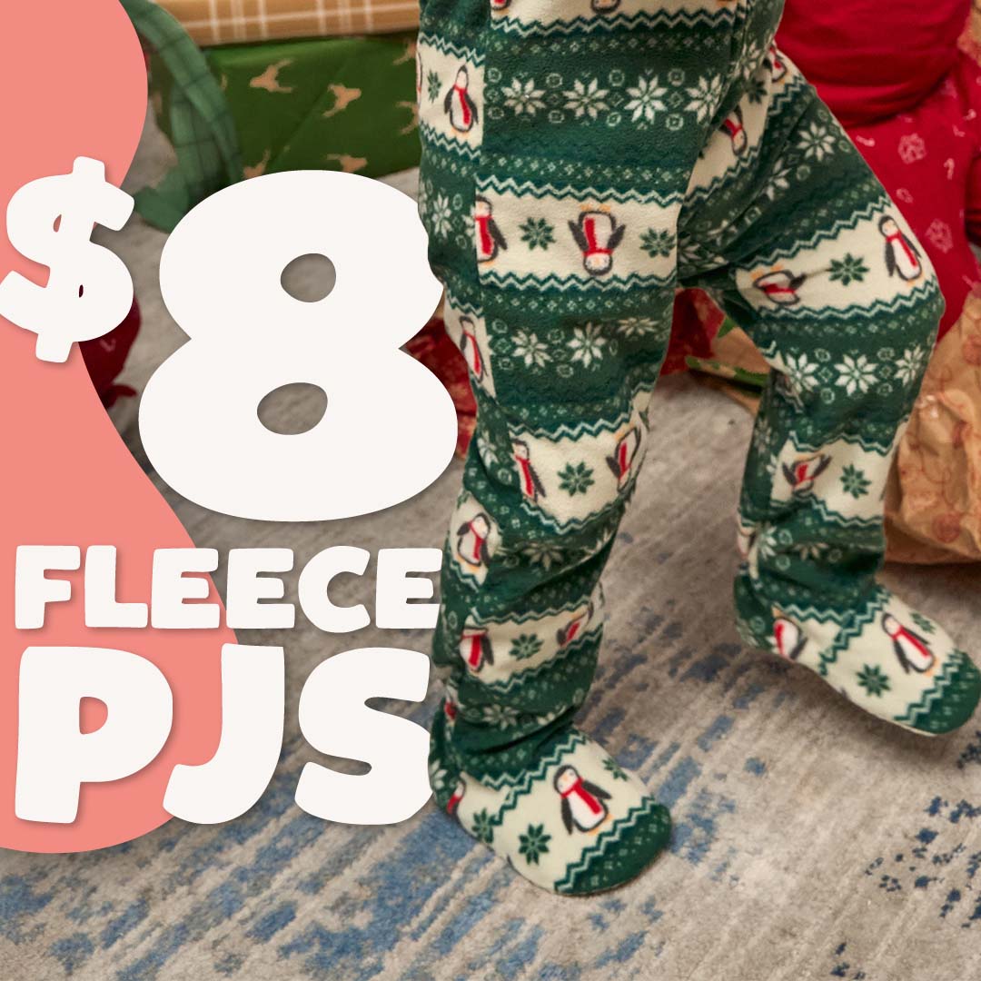 A close-up of a person wearing green and cream holiday fleece pajama pants with penguin and snowflake patterns, standing on a patterned rug with large promotional text overlay reading '$8 FLEECE PJS'.