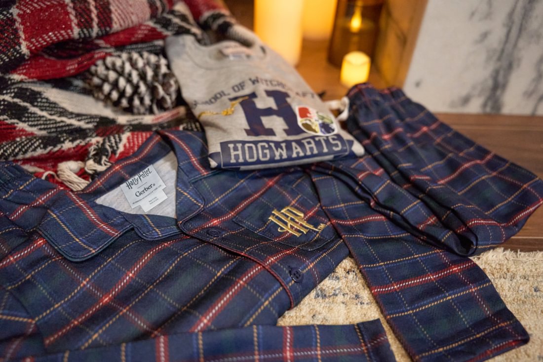 A cozy flat-lay of pajamas and a matching knit blanket on a wooden surface. The pajama set is navy blue plaid with red and thin yellow stripes, featuring a gold embroidered monogram on the chest.