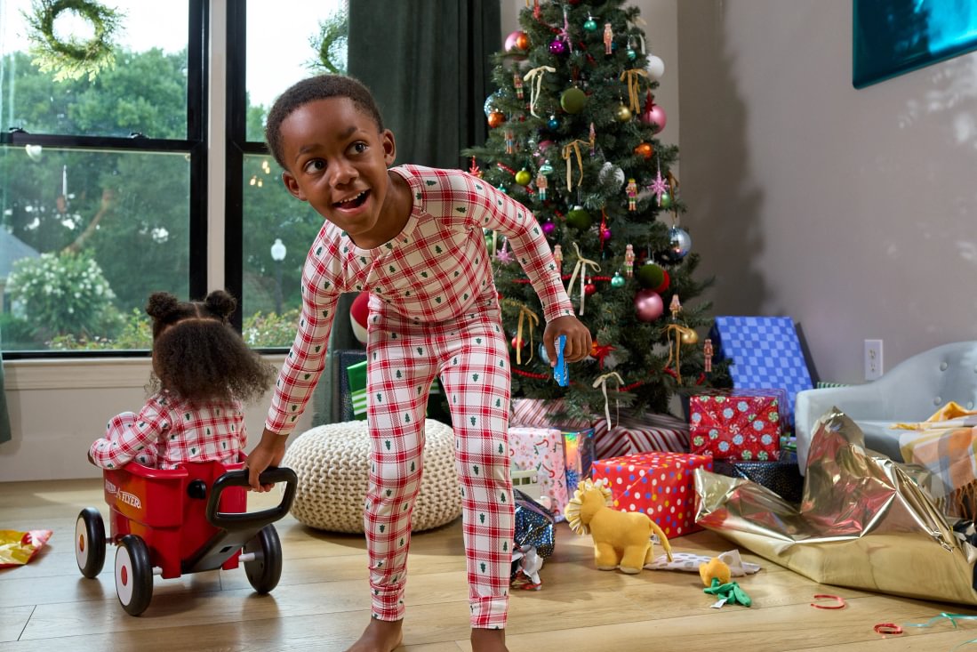 A child wearing red and white checkered pajamas stands in a living room near a Christmas tree and wrapped gifts, while another child sits in a red wagon nearby.