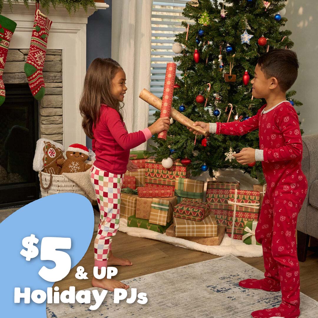 Two children in matching red holiday pajamas play with a rolled piece of red wrapping paper in a decorated living room with a Christmas tree, stockings, and wrapped gifts. A blue promotional badge in the corner reads '$5 & UP Holiday PJs'.