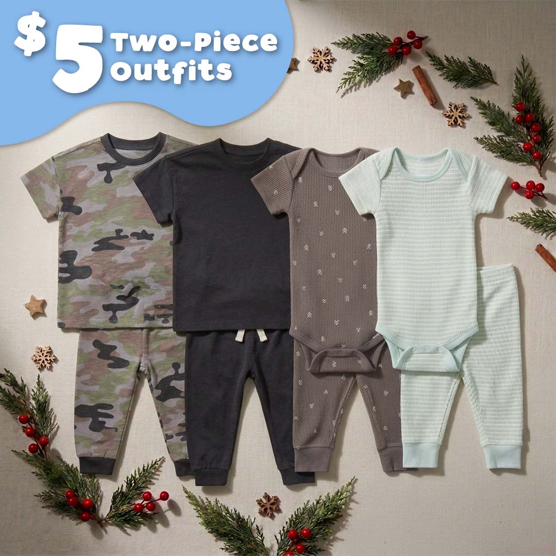 Four baby two-piece outfits arranged on a neutral fabric background with holiday greenery and red berries. The set includes a camouflage pajama set, a dark charcoal long-sleeve top with matching pants, a taupe bodysuit with small star print and matching pants, a pale mint striped bodysuit, and matching striped pants.