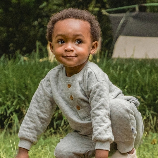 A toddler wearing a light gray long-sleeve button-front romper and matching pants, crouching outdoors on grass with trees in the background.
