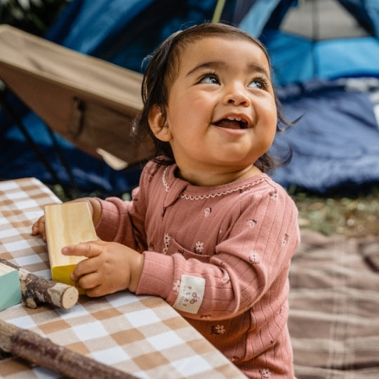 A smiling toddler wearing a dusty rose long-sleeve romper with small white floral prints, sitting at a checkered table outdoors near a tent.