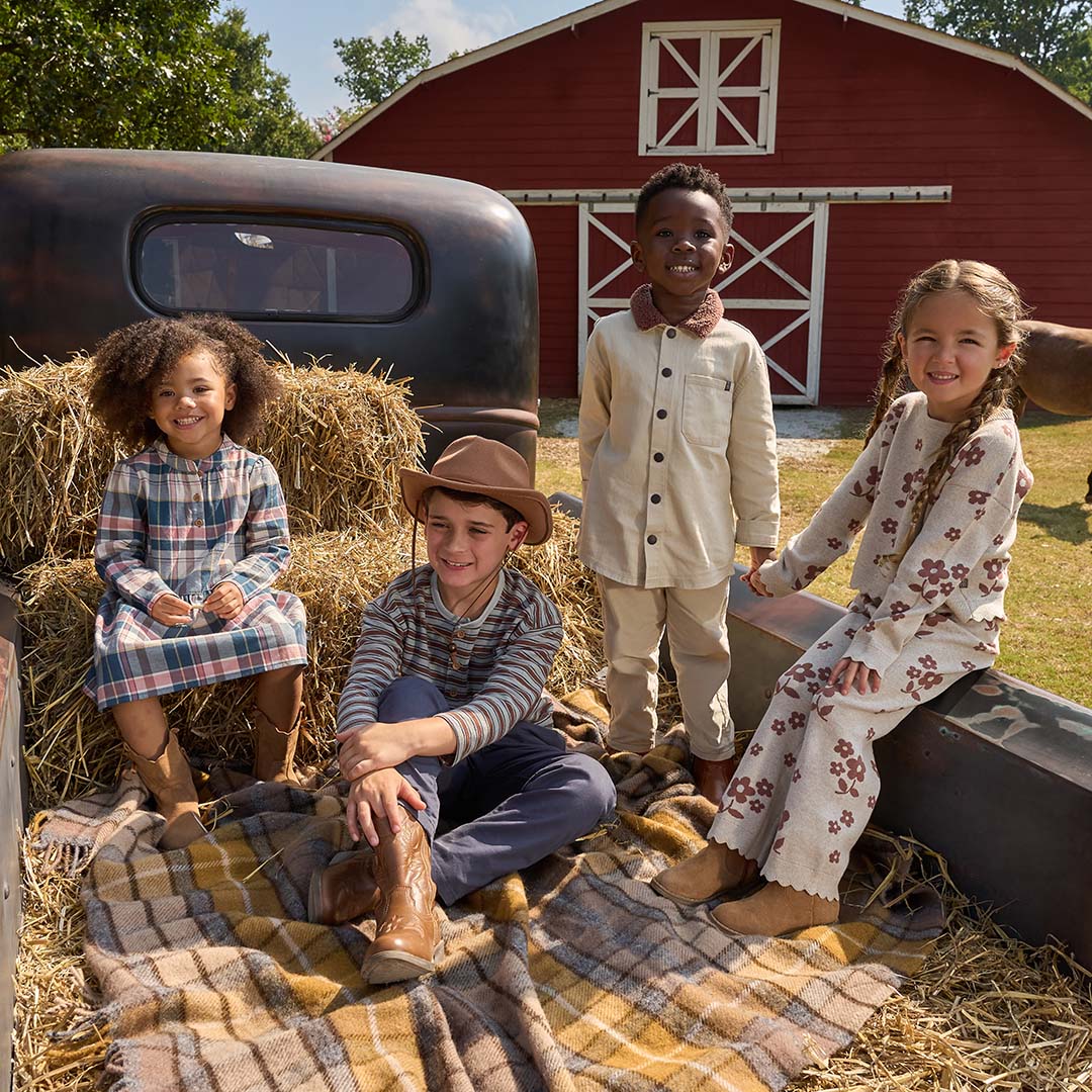 Four children dressed in rustic western and country-style clothing pose together in the bed of a vintage pickup truck on a straw blanket, with a red barn and trees in the background.