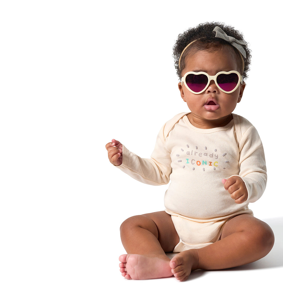 A baby wearing a cream long-sleeve bodysuit with 'already ICONIC' printed on the chest, heart-shaped sunglasses, and a gray bow headband, sitting against a white background.