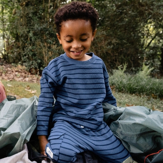 A young child wearing a navy blue long-sleeve waffle-knit pajama set with thin horizontal stripes, sitting outdoors on a green camping tarp with a forest background.