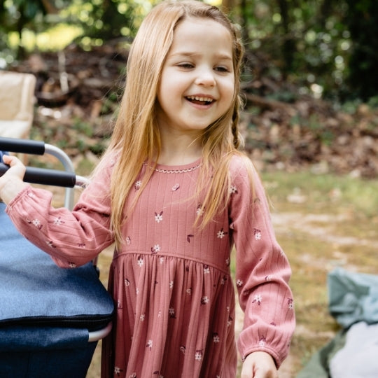A young girl with long blonde hair smiles while wearing a dusty rose long-sleeve dress with small white floral prints, standing outdoors next to a blue stroller.