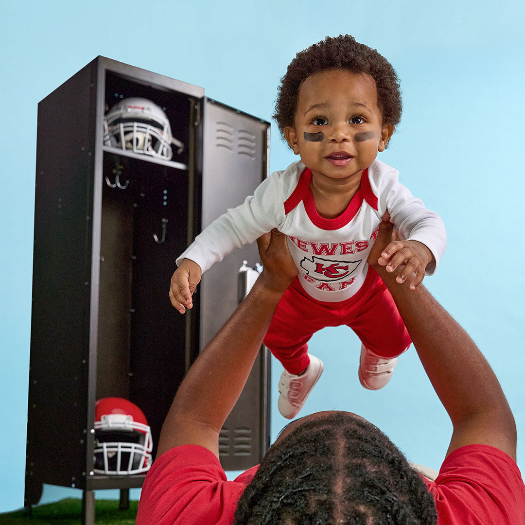 A baby wearing a red and white Kansas City Chiefs raglan shirt and matching pants is being held up by an adult in a red shirt, with a black football locker and helmets in the background against a light blue backdrop.