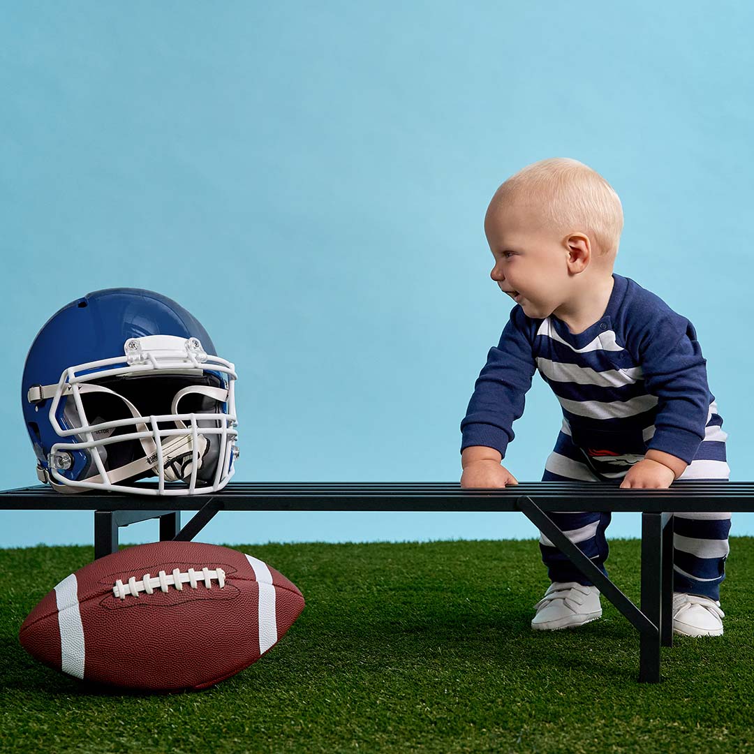 A baby in a navy and white striped long-sleeve romper and matching pants stands beside a black bench on artificial grass, looking at a blue football helmet and a brown football. The background is a solid light blue studio backdrop.