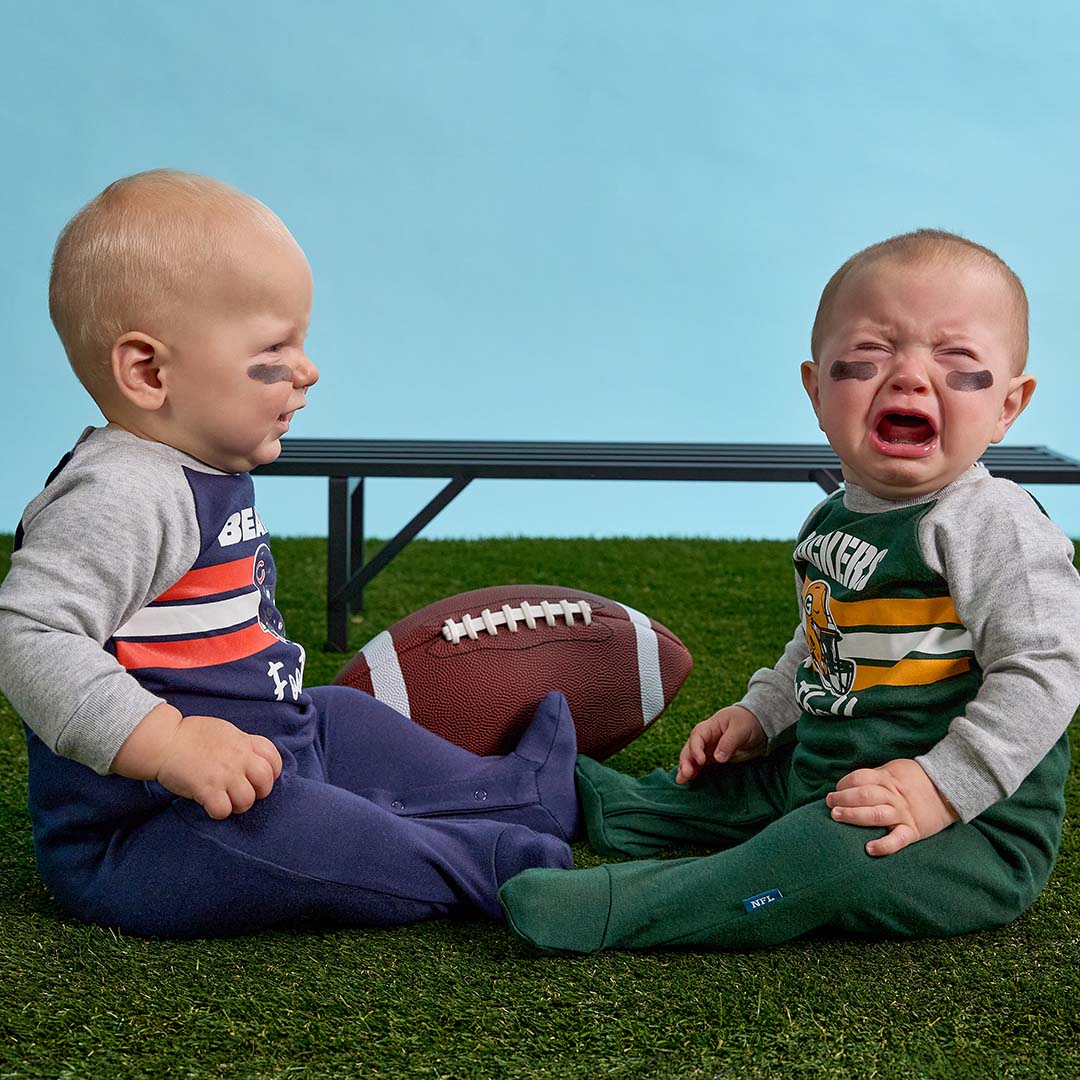 Two babies sitting on artificial grass wearing matching NFL team pajamas, with a football between them. The baby on the left wears a navy, red, and white outfit, and the baby on the right wears a green, yellow, and white Green Bay Packers outfit.
