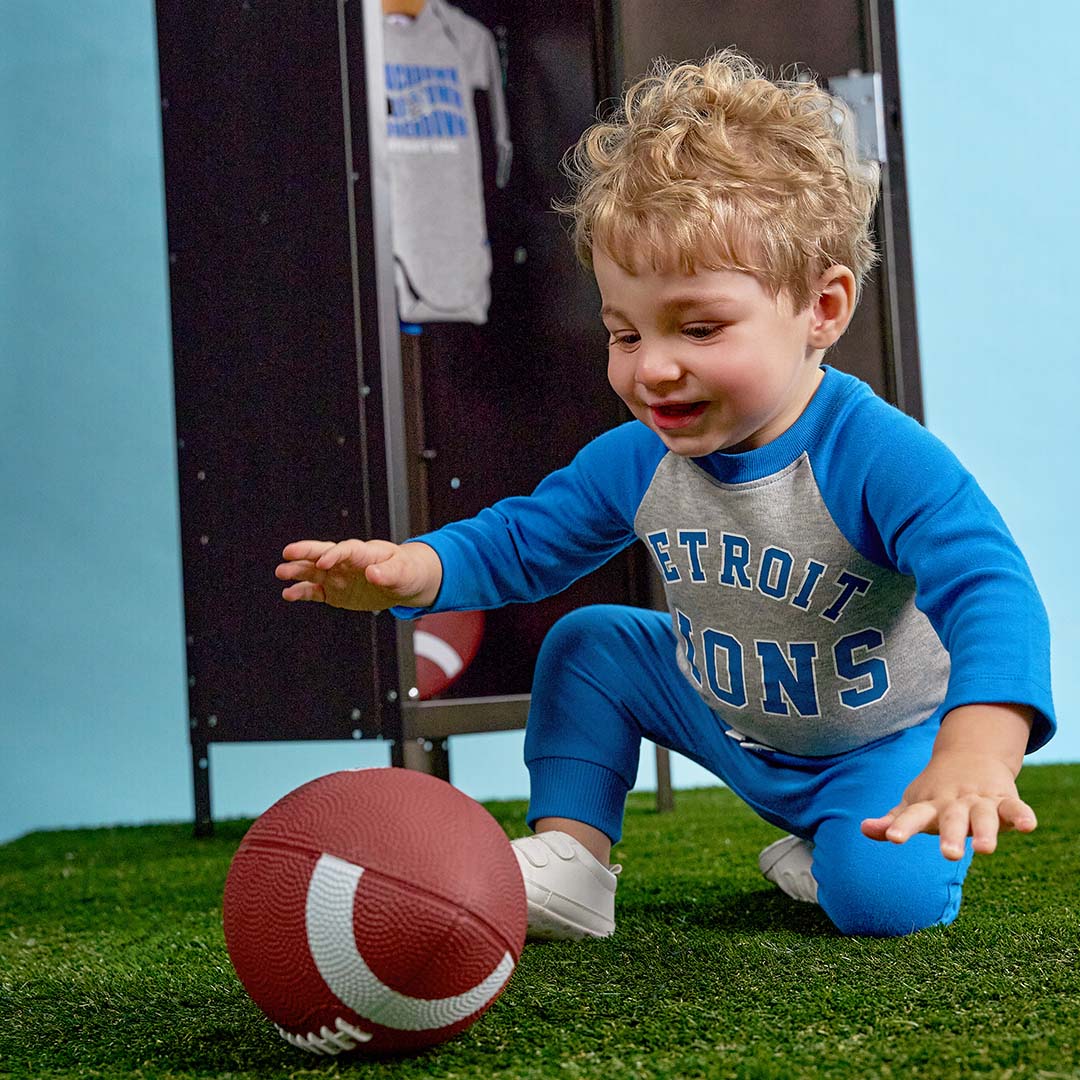 A toddler wearing a blue and gray Detroit Lions long-sleeve pajama set kneels on artificial turf next to a football, with a locker and another child visible in the background.
