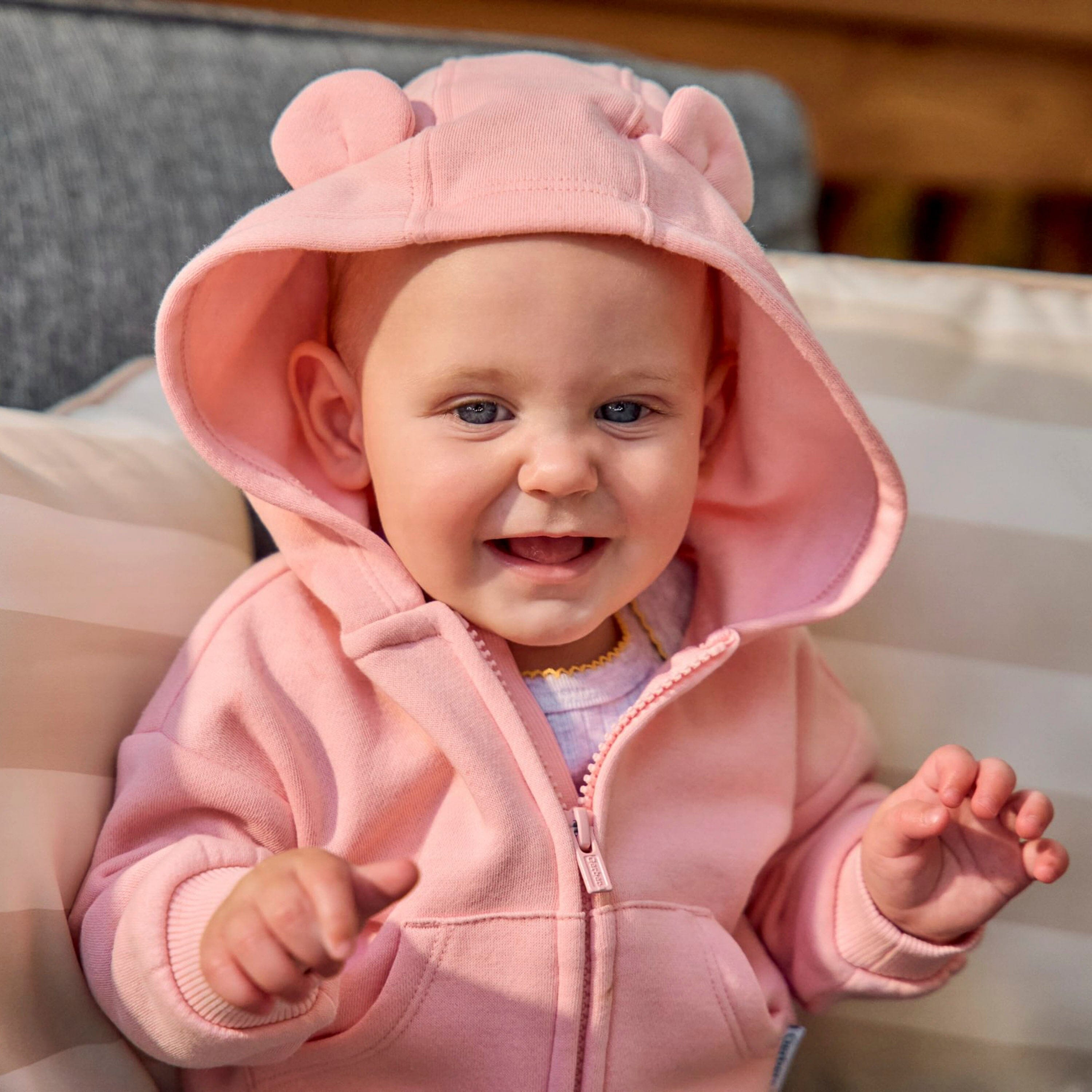 A baby wearing a pink zip-up hooded sweatshirt with bear ears on the hood, sitting on a striped cushion.