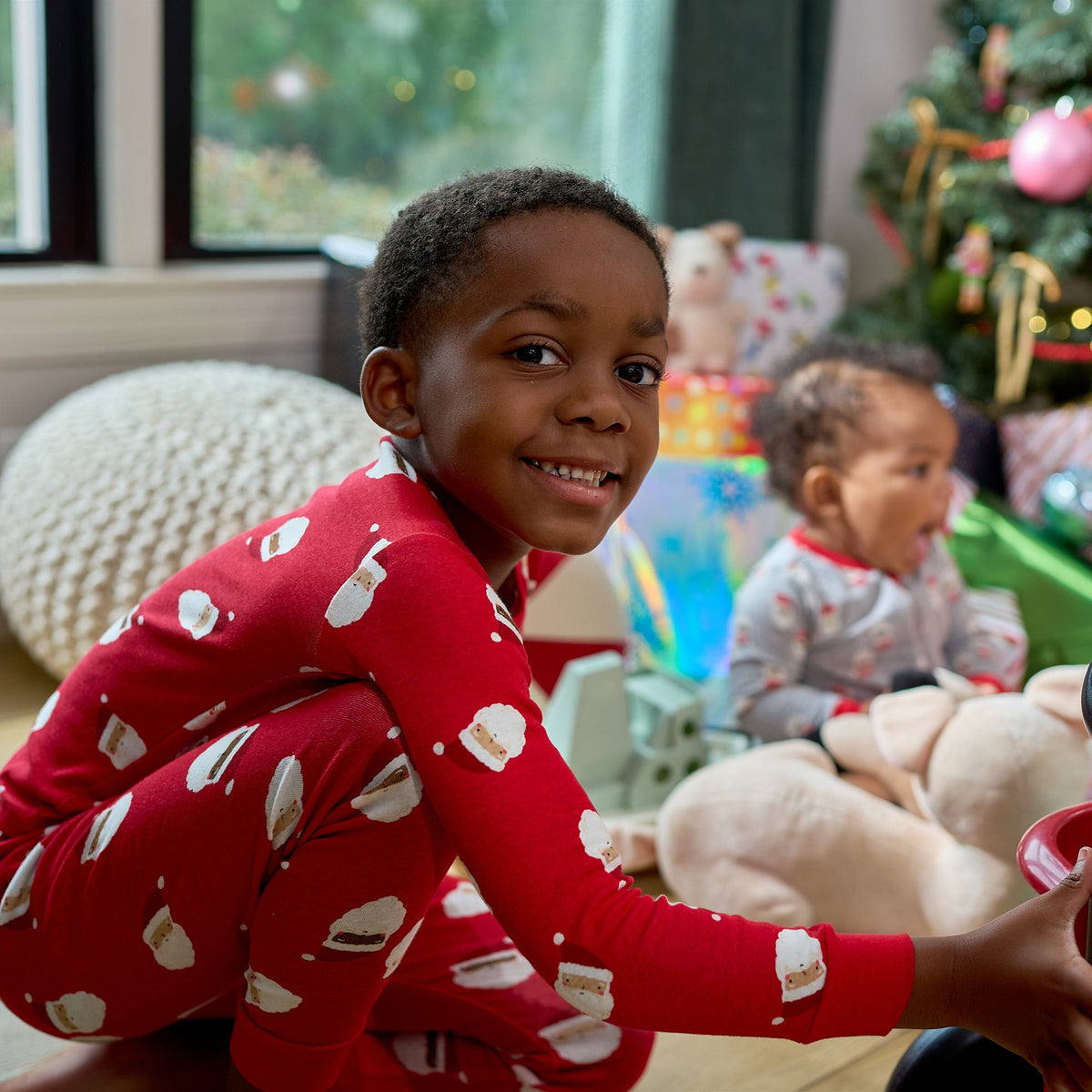 A young boy wearing red Christmas-themed pajamas with white cookie prints sits smiling in a living room, with a baby in matching pajamas and a decorated Christmas tree in the background.