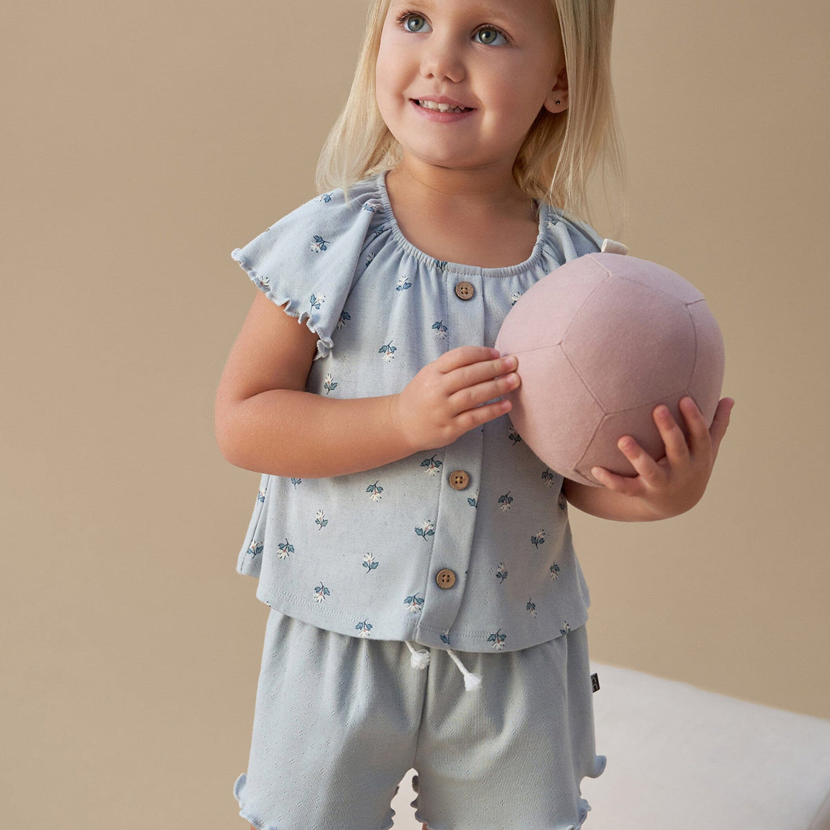 Young girl holding a pink ball wearing a light blue outfit with ruffled sleeves against a beige background