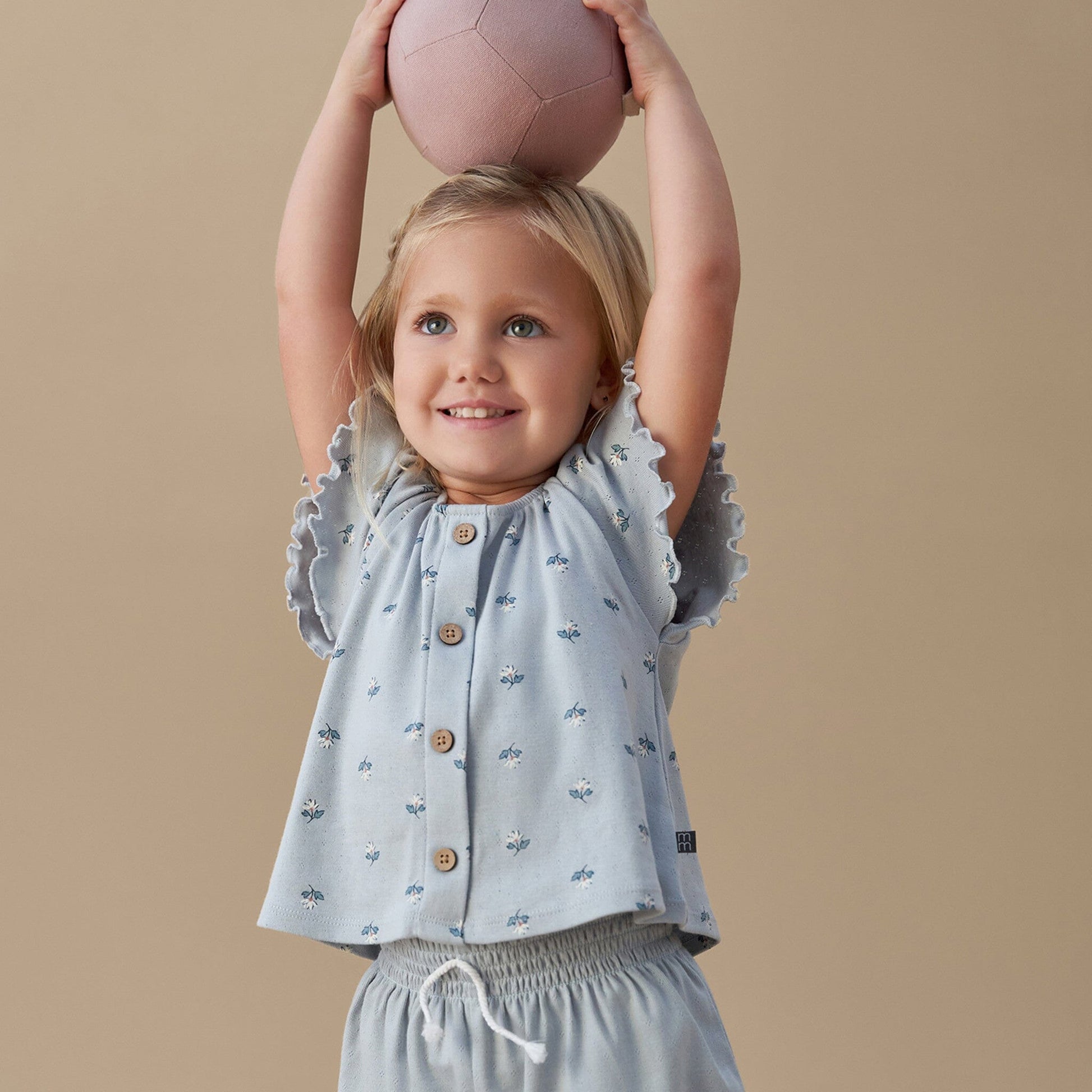 Child wearing a light blue top with ruffled sleeves and patterned pants, holding a pink ball above their head against a beige background.