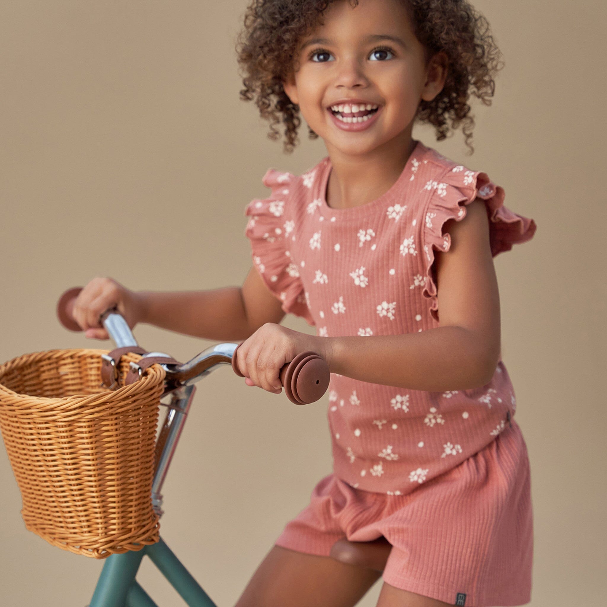 Child in a pink floral outfit riding a bicycle with a basket on a beige background