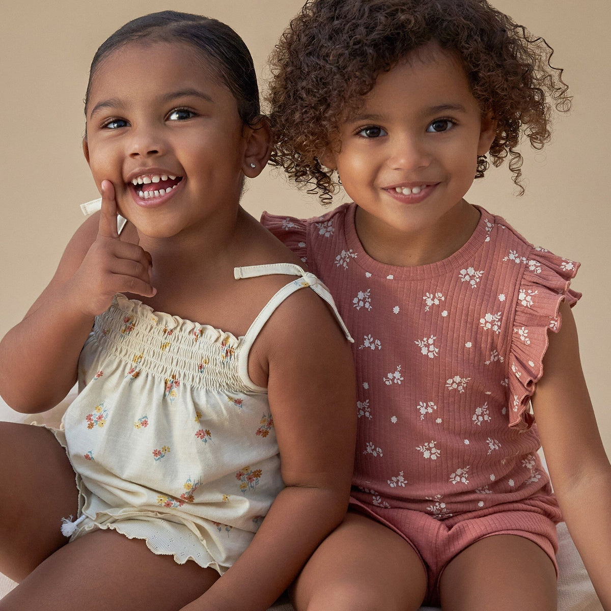 Two young girls sitting together wearing floral outfits on a beige background